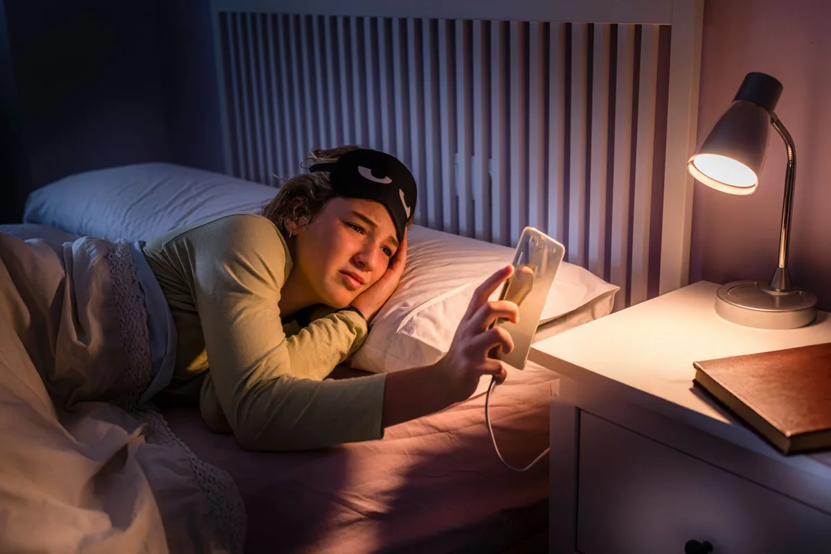 A young woman lying in bed at night, lit by her phone screen, watching for a reply that has not come
