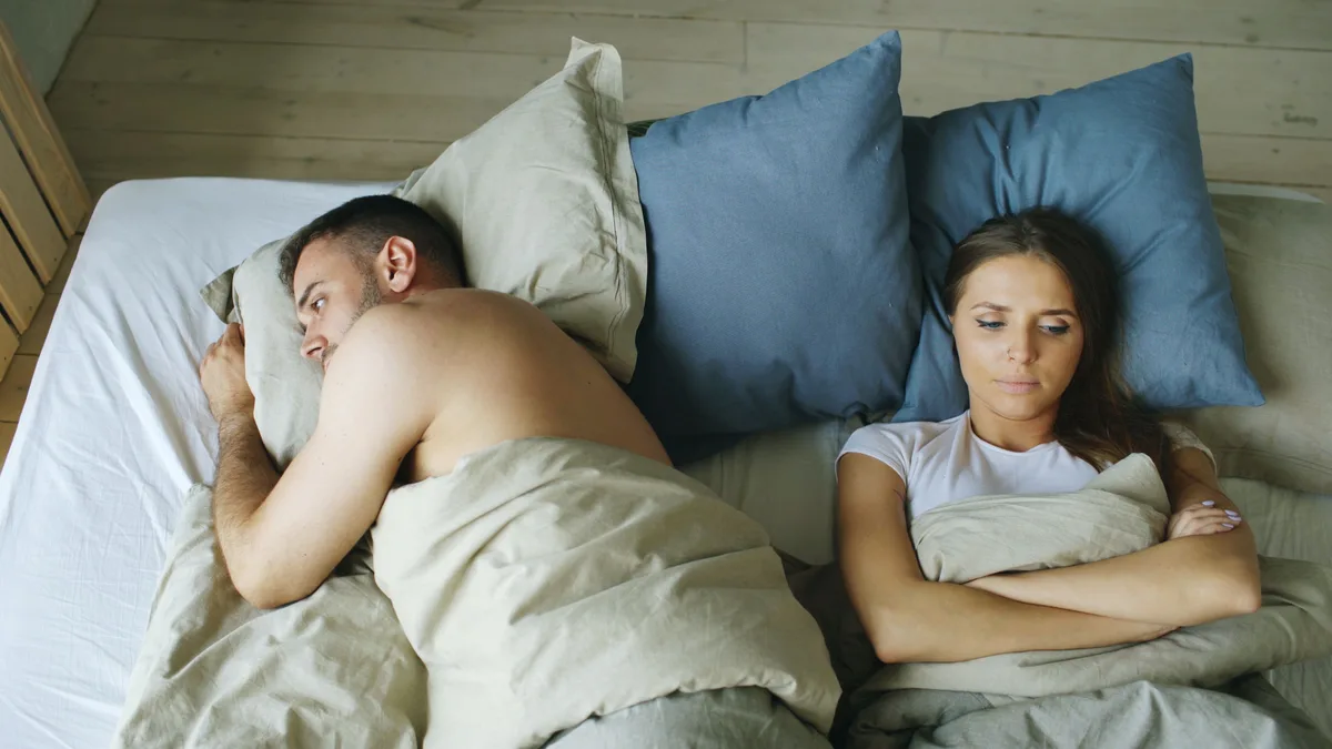 A couple lying awake in bed, turned away from each other with a gap between them