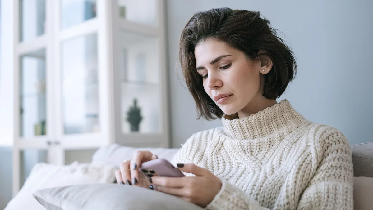 A woman sitting on a sofa, looking down at her phone with a tense, uncertain expression