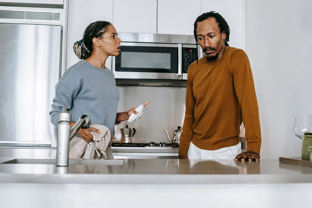 A woman gesturing with a plate mid-argument in a kitchen while her partner looks down in silence